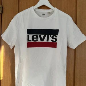 Vit t-shirt från Levi's - Säljer en klassisk vit t-shirt från Levi's med deras ikoniska logga i svart och rött på bröstet. T-shirten har en normal passform och korta ärmar. Perfekt för en avslappnad stil. Hör av dig vid frågor