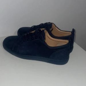 Mörkblå sneakers från Christian Louboutin - Snygga mörkblå sneakers i mocka från Christian Louboutin. Skorna har en klassisk design med snörning och en ikonisk röd sula. Perfekta för att ge din outfit en lyxig touch.