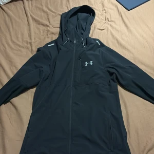 Svart jacka från Under Armour - Säljer en svart jacka från Under Armour med huva och dragkedja. Jackan har långa ärmar och är perfekt för träning eller vardagsbruk. Den är tillverkad i ett syntetmaterial som ger bra skydd mot väder och vind. Pris kan diskuteras