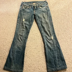 Blå bootcut jeans från True Religion - Säljer ett par snygga blå bootcut jeans från True Religion. De har en klassisk femficksdesign och slitna detaljer på framsidan. Tillverkade i USA med en cool vintage-look. Perfekta för dig som vill ha en avslappnad men stilren stil. 🕶️