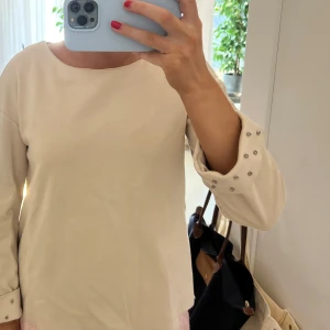 Beige långärmad tröja med pärldetaljer - Säljer en stilren beige tröja med långa ärmar och dekorativa pärlor på ärmsluten. Perfekt för en elegant look. Tröjan har en enkel design med rund hals och är tillverkad i ett mjukt material. Tröjan har tappat två stycken stenar som satt på slutet av ärmen! Den är i storlek XS men passar också S!💗