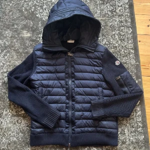 Cardigan Moncler - Säljer en snygg mörkblå jacka från Moncler med quiltad design och stickade ärmar. Jackan har en praktisk huva och dragkedja framtill. Perfekt för kyligare dagar!