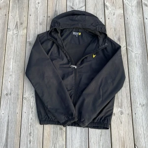 Svart vindjacka från Lyle & Scott - Säljer en svart vindjacka från Lyle & Scott med huva och dragkedja. Jackan har ett gult logotypbroderi på bröstet och elastiska muddar. Perfekt för blåsiga dagar! 🌬️