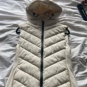Beige dunväst med huva - Snygg beige dunväst med dragkedja och huva. Perfekt för kyliga dagar. Västen har ett quiltat mönster och är både stilren och praktisk.