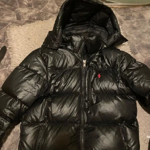 Svart dunjacka från Polo Ralph Lauren - Säljer en snygg svart dunjacka från Polo Ralph Lauren. Jackan har en huva och dragkedja framtill. Den är perfekt för kalla dagar och har en klassisk design med det ikoniska röda logotypen på bröstet.Inköpt för 4500 kronor. Hängremsan (finns i andra bilden) har släppt minimalt, men går fortfarande utmärkt att hänga jackan på en krok.