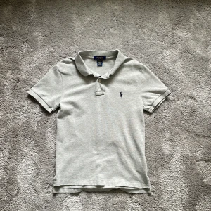 Grå pikétröja från Ralph Lauren - Säljer min sons grå pikétröja från Ralph Lauren med klassisk krage och knappdetalj. Tröjan har en broderad logotyp på bröstet och är kortärmad. Perfekt för en avslappnad men ändå snygg look.