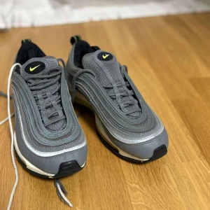 Grå Nike Air Max 97 - Snygga grå Nike Air Max 97 med ikonisk design och synlig Air-enhet i sulan. Skorna har en stilren grå färg med gula detaljer och snörning. Perfekta för både stil och komfort.