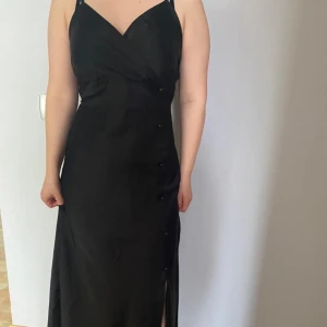 Svart långklänning med knappar - Elegant svart långklänning med knappar längs sidan och en subtil slits. Klänningen har smala axelband och en omlott-design framtill. Perfekt för en stilren look.