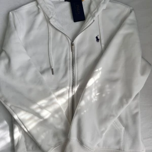 Vit zip från Ralph Lauren - Säljer en stilren vit hoodie från Ralph Lauren med dragkedja och broderad logga i mörkblått på bröstet. Tröjan har långa ärmar och en praktisk huva med snörning. Perfekt för en avslappnad look!