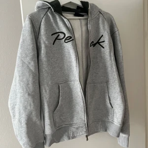 Grå hoodie från Peak Performance - Säljer en grå hoodie från Peak Performance med dragkedja och svart text på framsidan. Perfekt för en avslappnad stil. Storlek L men passar M-S