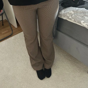Bruna rutiga byxor - Snygga bruna kostymbyxor med ett diskret rutigt mönster. Byxorna har en tight passform och är högmidjade, vilket ger en smickrande siluett. Perfekta för en stilren look.