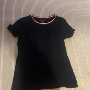 Svart t-shirt från Tommy Hilfiger - Snygg svart t-shirt från Tommy Hilfiger med korta ärmar och en stilren design. Kragen har en röd, vit och blå randig detalj som ger en sportig touch. Perfekt för en casual look!
