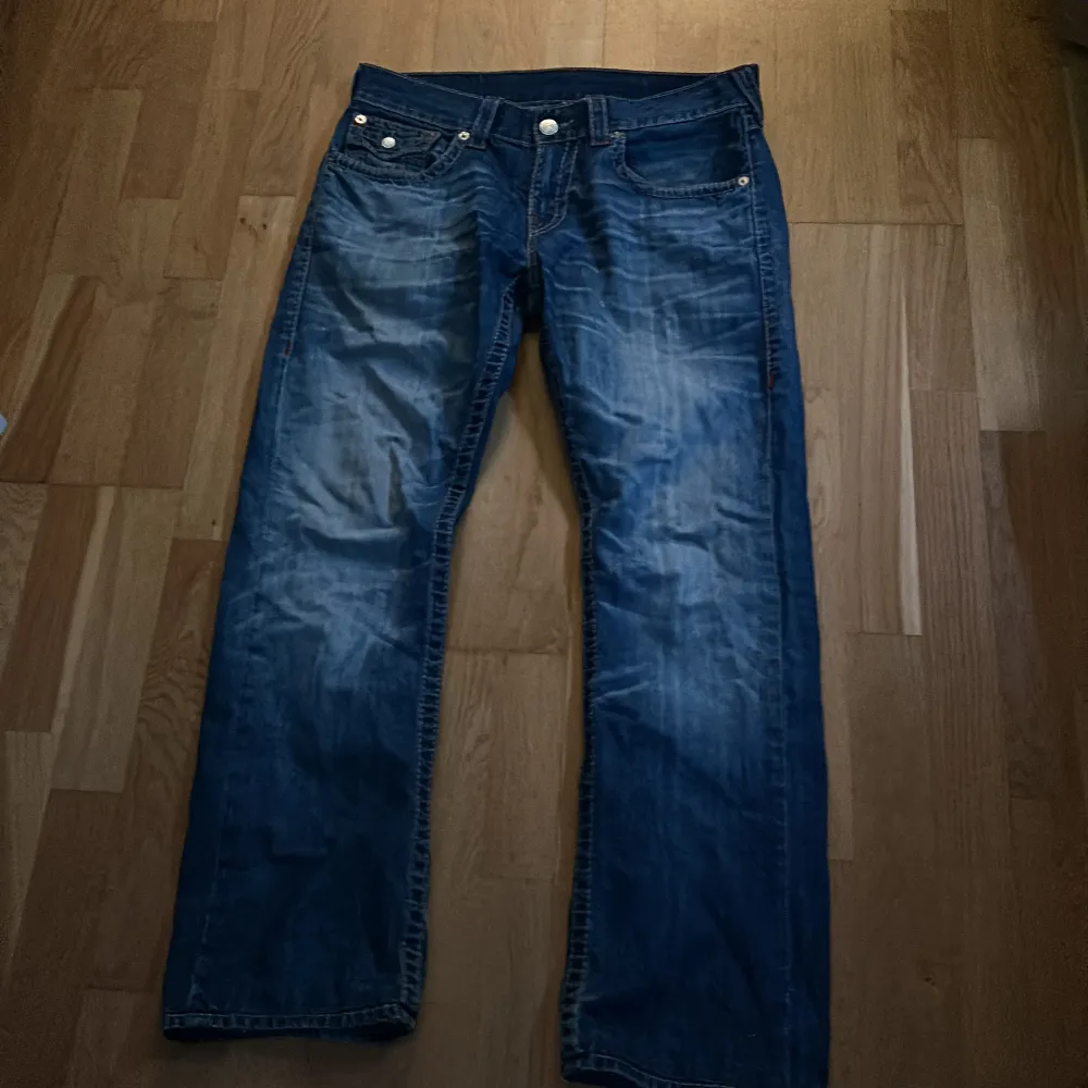 Säljer ett par snygga blå jeans från True Religion med klassisk femficksdesign och knappar. Jeansen har en straight passform och är tillverkade i slitstarkt denim. Perfekta för en avslappnad stil.. Farkut & Housut.