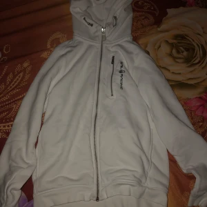 Vit hoodie från Sail Racing - Säljer en stilren vit hoodie från Sail Racing med dragkedja och justerbar huva. Perfekt för kyliga dagar. Den har en praktisk bröstficka med dragkedja och logotyp på bröstet.