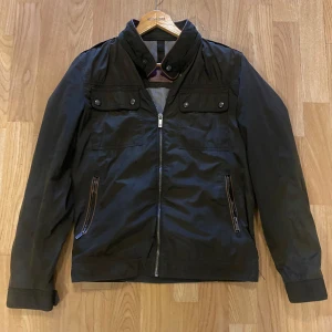 Svart jacka från Massimo Dutti - Snygg svart jacka från Massimo Dutti med dragkedja och knappar. Jackan har långa ärmar och två fickor med dragkedja framtill. Perfekt för en stilren look. Pris är ej hugget i sten!