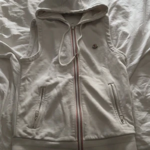 Moncler hoodedwest - Säljer en stilren vit väst från Moncler. Alldeles för liten därför jag säljer den. Den är äkta och är storlek S men väldigt liten i storlek. Den är köpt på kaspersheat för 1800. Finns ett mini hål fram på tröjan som man knappt ser som ni ser på bild 2