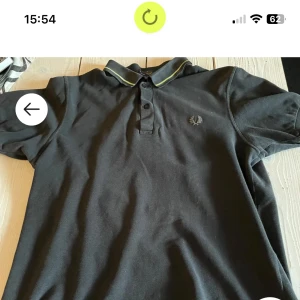 Svart pikétröja från Fred Perry - Säljer en klassisk svart pikétröja från Fred Perry med grön rand på kragen. Tröjan har korta ärmar och är tillverkad i 100% bomull. Den har en broderad logotyp på bröstet. Perfekt för en stilren look!