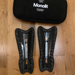 Svarta benskydd från Monolit - Säljer ett par svarta benskydd från Monolit. De har en modern design med en slät yta och är lätta att bära. Perfekta för fotbollsspelare som söker både skydd och stil. Levereras med en praktisk förvaringsväska. Ser helt nya ut och vöger typ ingenting jag gav 800kr för dessa dem tvätts innan dem levereras.