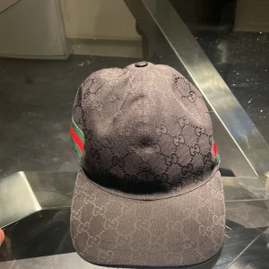 Svart keps med mönster - Snygg svart keps med diskret mönster och justerbart spänne baktill. Kepsen har en grön och röd randdetalj på sidan som ger en extra touch. Perfekt för en stilren look.