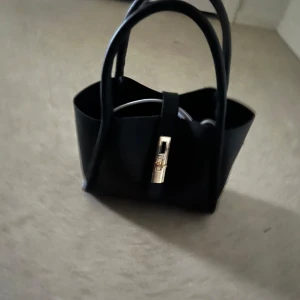 Svart handväska med gulddetalj - Elegant svart handväska med stilren gulddetalj på framsidan. Perfekt för att ge en touch av lyx till din outfit. Väskan har två handtag för enkel bärning. Rymmer väldigt mycket väldigt fint skick inget sönder inga fläckar 