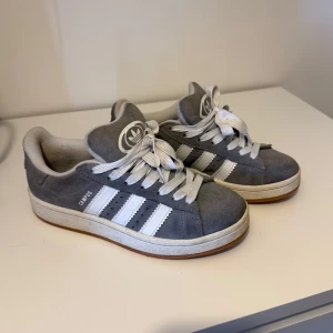Grå Adidas Campus sneakers - Snygga grå Adidas Campus sneakers med vita ränder och snörning. Skorna har en klassisk design med gummisula och är perfekta för en avslappnad stil. De är tillverkade i mocka och har en bekväm passform.Pris kan definitivt diskuteras 