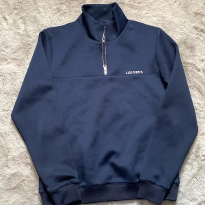 Mörkblå tröja med dragkedja från Les Deux - Säljer en stilren mörkblå tröja från Les Deux med halv dragkedja och broderad logga på bröstet. Perfekt för en avslappnad men snygg look. Tröjan har långa ärmar och ribbade muddar för extra komfort. Pris går att diskuteras vid snabb affär