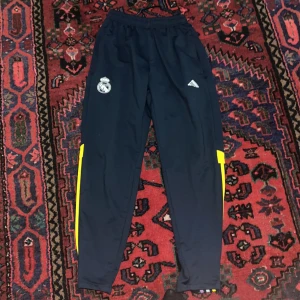 Träningsbyxor från Adidas - Snygga träningsbyxor från Adidas i mörkblått med gula och flerfärgade detaljer längs sidorna. Byxorna har elastisk midja och broderad logga. Perfekta för sport och aktivitet.