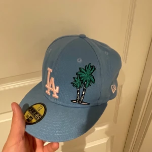 Ljusblå LA keps från New Era 59FIFTY - Säljer en ljusblå keps från New Era 59FIFTY med LA-logga i vitt och broderade palmer på sidan. Kepsen har även en broderad patch på sidan och är tillverkad i 100% polyester. Klassisk flat peak-modell. Använts 2 gånger
