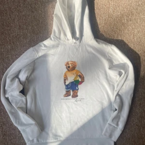 Vit hoodie med Polo Bear från Ralph Lauren - Vit hoodie från Ralph Lauren med ikoniska Polo Bear-trycket framtill. Tröjan har huva och långa ärmar, samt mjukt material som känns skönt mot huden. Perfekt för en avslappnad och stilren look. Skick: 8/10 bra fast köpt secondhand. Priset är diskuterbart, skriv för mer info.