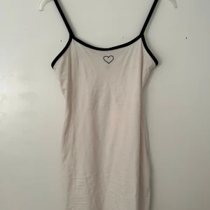 Vit kortklänning med svarta detaljer och hjärta - Söt vit kortklänning med svarta tunna axelband och svarta kanter. Liten broderad hjärt-detalj framtill. Perfekt för varma dagar eller som ett lagerplagg. Enkel och stilren design.