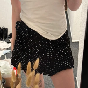 Svart prickig kortkjol med volang och knyt - Säljer en svart kortkjol med vita prickar och volangkant. Kjolen har ett knytband i midjan och omlottkänsla, vilket ger en söt och trendig look. Perfekt för dig som gillar lekfulla detaljer och mönster.