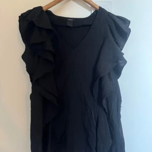 Svart volangtopp från Lindex - Säljer en svart topp från Lindex med volangdetaljer längs axlar och framstycke. Toppen har v-ringning och är ärmlös! Storlek S