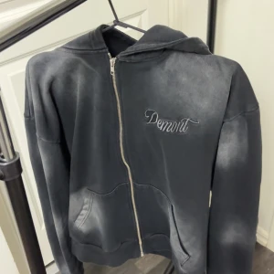 Svart hoodie med dragkedja från Demont - Cool svart hoodie från Demont med dragkedja framtill och broderad logga på bröstet. Tröjan har huva och två fickor framtill. Perfekt för en avslappnad stil.