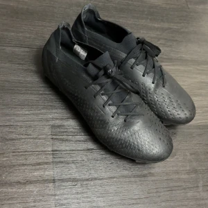 Svarta fotbollsskor från Adidas - Säljer ett par helsvarta fotbollsskor från Adidas med snörning och låg profil. Skorna har en texturerad ovandel och platt sula anpassad för spel på konstgräs eller gräs. Perfekta för dig som vill ha en stilren look på planen.