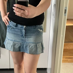 Jeanskjol med volangkant - Säljer en blå jeanskjol med volangdetalj längst ner. Kjolen har klassiska fickor och bälteshällor och stängs med knapp och dragkedja. Perfekt för dig som gillar en kort och trendig look.
