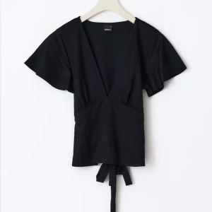 Svart omlottopp med knytband - Snygg svart omlottopp med v-ringning och korta ärmar. Toppen har knytband i midjan som ger en fin passform och en croppad look. Perfekt att styla med både jeans och kjol. Den har ett litet hål men inget man tänker på 