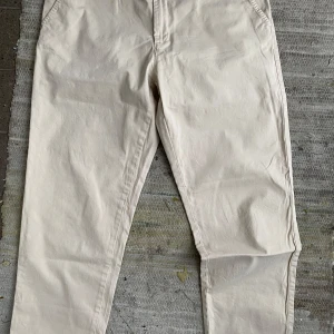 Beige chinos från Holly and Whyte - Snygga beige chinos från Holly and Whyte med klassisk passform. Byxorna har framfickor, bakfickor med knappar och en liten metallogo ovanför ena bakfickan. Perfekta för en stilren look.