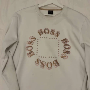 Vit sweatshirt från Hugo Boss - Vit sweatshirt med rund hals från Hugo Boss. Tryck med ordet 'BOSS' i rött och beige i cirkel på bröstet samt små 'HUGO BOSS' i mitten. Långärmad och i mjukt material, perfekt för en avslappnad stil.