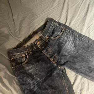 Mörkblå jeans från Nudie Jeans - Säljer ett par mörkblå jeans från Nudie Jeans med klassisk femficksdesign och orangea sömmar. Jeansen har raka ben och en snygg slitning framtill samt Nudie-logga i läder baktill i midjan.