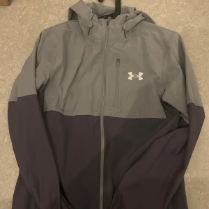 Grå och svart vindjacka från Under Armour - Snygg tvåfärgad vindjacka från Under Armour i grått och svart. Jackan har huva, dragkedja framtill och en diskret logga på bröstet. Perfekt för träning eller när du vill ha en sportig look.