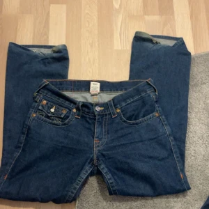 Blå jeansbyxor från True Religion - Säljer ett par klassiska blå jeans från True Religion med ikoniska bakfickor och kontrastsömmar. Byxorna har normal passform och raka ben, perfekt för en avslappnad stil. Klassisk femficksmodell och knappgylf.