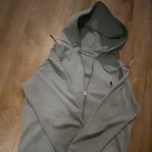 Polo Ralph Lauren - Grå hoodie med dragkedja från Polo Ralph Lauren. Klassisk design med huva och snörning, samt den ikoniska broderade loggan på bröstet. Perfekt för en avslappnad stil.Ny pris ligger på 2200.Dm för mer bilder.Ingen hål på hoodie eller nåt 