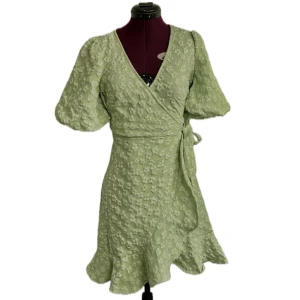 Ljusgrön blommig omlottklänning - Superfin ljusgrön omlottklänning med blommigt mönster och volangdetaljer nertill. Klänningen har puffärmar och knyts i midjan för en smickrande passform. Perfekt för vår och sommar! Endast använd en gång! (Ca 85cm lång, 65cm midja, 85cm byst)