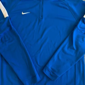 Blå långärmad träningströja från Nike - Säljer en blå långärmad träningströja från Nike med halv dragkedja och vit logga på bröstet. Tröjan är tillverkad i Dri-Fit material som andas och passar perfekt till sport och träning. Snygg och sportig modell med bekväm passform.