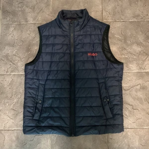 Hugo boss väst - Snygg mörkblå dunväst från Hugo med diskret quiltad design och röd logotyp på bröstet. Västen har dragkedja framtill och två fickor med knappstängning. Perfekt för lager-på-lager och stilren look. Bra skick