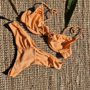Sunset orange bikini från Nelly - Bikinitopp från Nelly, ovadderad kupa med bygel. Smala, justerbara axelband och knytband mellan byst. Helfodrad bikinitrosa med scrunch och stringmodell.  Överdelen är i storlek 70B och trosan i S.