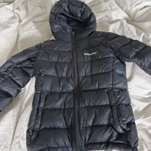 Svart dunjacka från Peak Performance - Säljer en svart dunjacka från Peak Performance med huva och dragkedja framtill. Jackan har quiltad design och är perfekt för kyliga dagar. Klassisk look med logga på bröstet och två fickor med dragkedja.