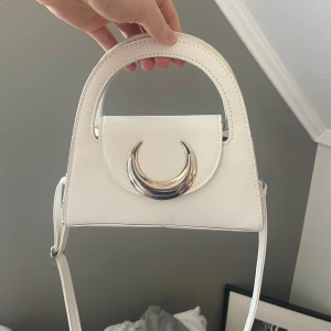 Vit axelväska med silverdetalj - Säljer en stilren vit axelväska med unik halvmåneformad silverdetalj på framsidan. Väskan har ett topphandtag och justerbar axelrem. Perfekt för att lyfta vilken outfit som helst med en modern touch.