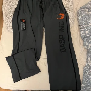 Mörkgrå träningsbyxor från GASP INC - Säljer ett par mörkgrå träningsbyxor från GASP INC med svarta detaljer och tryck på benet. Byxorna har resår i midjan och dragsko för justerbar passform. Perfekta för gymmet eller andra träningspass.