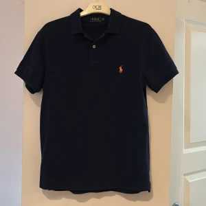 Mörkblå pikétröja från Polo Ralph Lauren - Klassisk mörkblå pikétröja från Polo Ralph Lauren med orange broderad logga på bröstet. Piken är i storlek M men passar också S. Perfekt nu till sommarn, hör av dig vid frågor/funderingar. Priset kan diskuteras vid snabb affär!